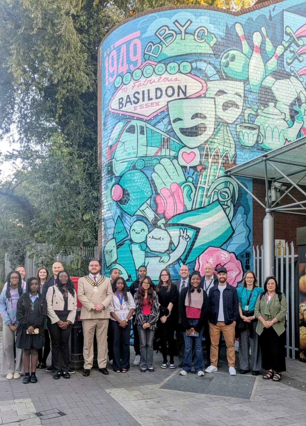 The full mural on the curved brick wall at Basildon station's entrance, in turquoise, blue and pink, showing a 'Welcome to Fabulous Basildon Essex' sign, 'Est 1949', the letters BBYC, theatre masks, a train, bowling pins and a teapot, with a group standing in front.