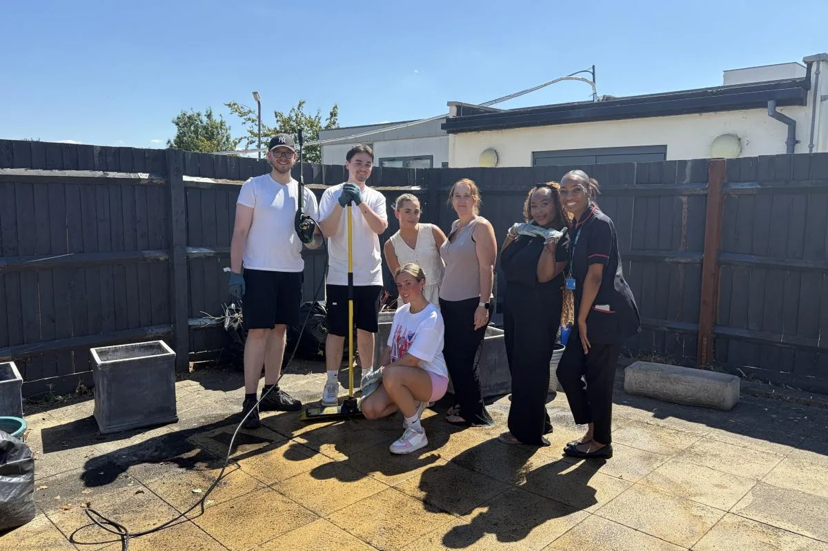 A group of volunteers in casual clothes and two hospital staff in uniform standing on a freshly jet-washed patio in front of a dark wooden fence, with a pressure washer and broom.